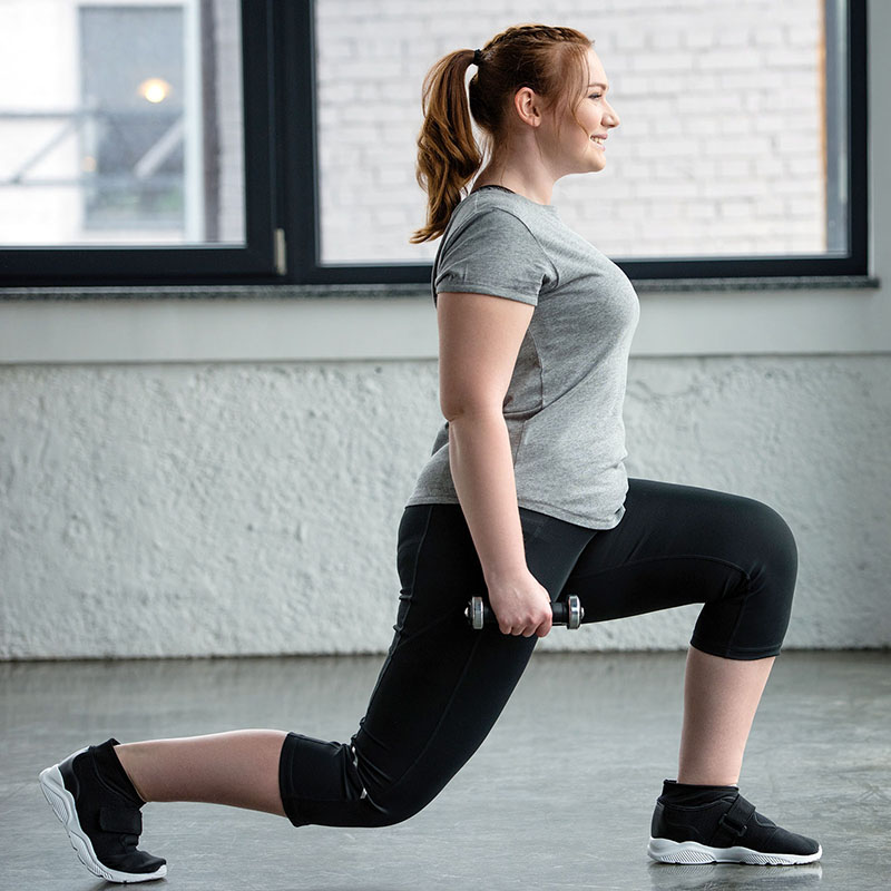 Obese girl performing lunges with dumbbell in gym